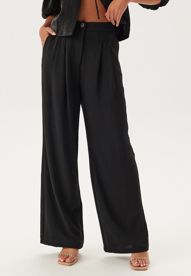 Pantalons larges noirs en tissu léger, avec des plis, une fermeture à bouton et une texture lisse. Associés à des sandales beiges.