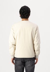 Beige, lett jakke med enkelt design, ribbede mansjetter og rund hals, vist fra baksiden mot en ensfarget bakgrunn.