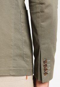 Kurtka oliwkowo-zielona o teksturowanym wykończeniu, z boczną kieszenią i czterema brązowymi guzikami na mankiecie z kontrastowym przeszyciem.