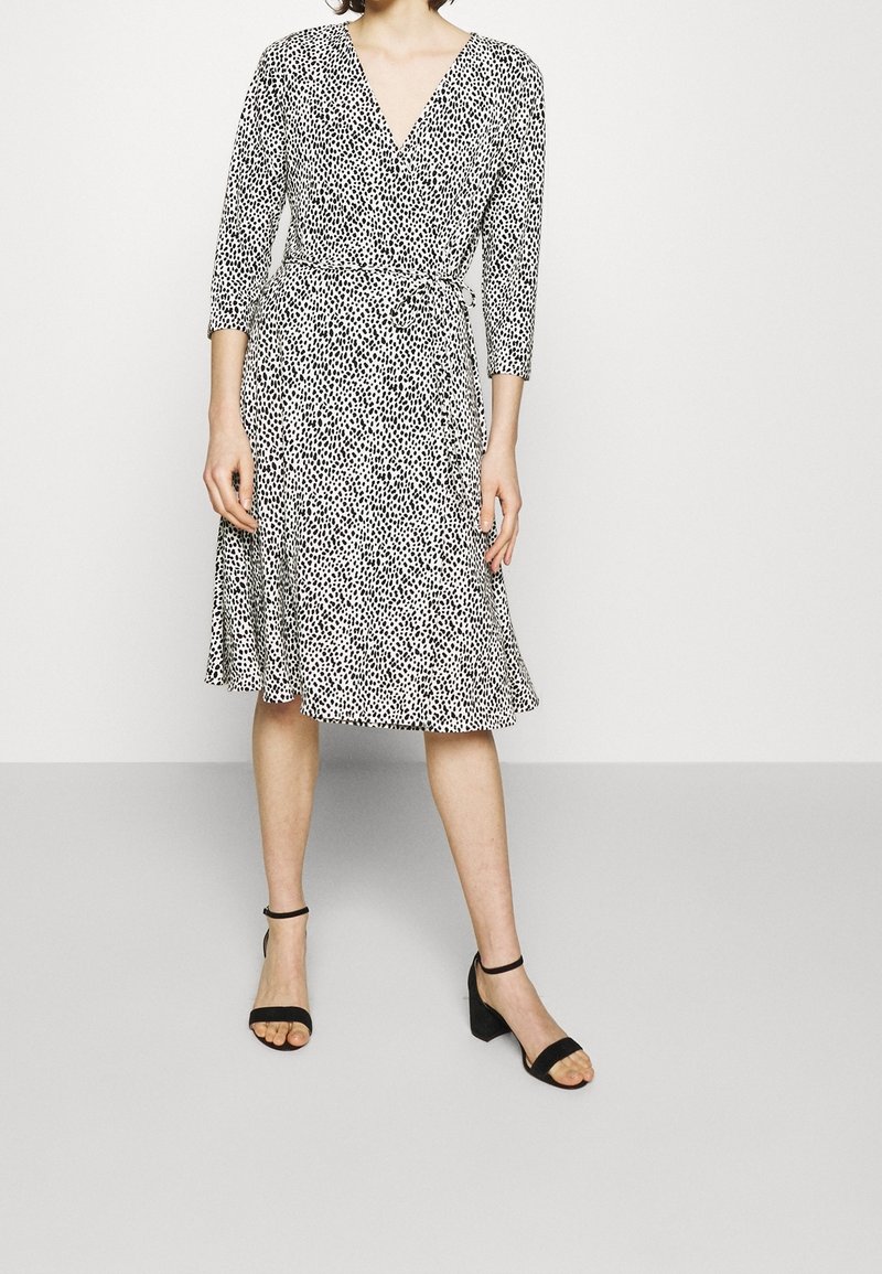 Schwarz-weißer Pünktchen-Wickelkleid mit Dreiviertelärmeln, V-Ausschnitt und gebundenem Taillenband, mit schwingendem Rock und glatter Textur.
