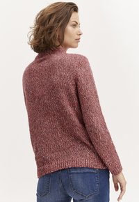 Adīta džemperis maigā, teksturētā rozā krāsā ar augstu apkakli un brīvu piegriezumu, valkāta ar zilām džinsiem, radot ikdienišķu izskatu.