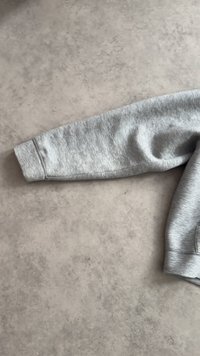 Rechter Ärmel eines hellgrauen Sweatshirts mit geripptem Bündchen, flach liegend auf einer strukturierten, hellen Oberfläche.