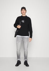 Schwarzer Pullover mit Logo, graue zerrissene Jeans, schwarze Sneakers und eine schwarze Mütze. Das Model trägt eine schwarze Schultertasche.