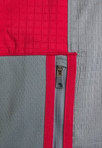 Gros plan sur un tissu rouge et gris avec une fermeture éclair verticale grise bordée de coutures rouges.