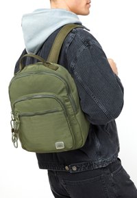 Maslinasto zelena ruksak s teksturiranim materijalom, više zip zatvarača, i remenom od mrežice. Mali logo prikazan sprijeda.