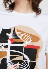 T-shirt en coton blanc présentant un motif graphique abstrait en noir, rouge, marron et beige, avec des formes circulaires et angulaires.