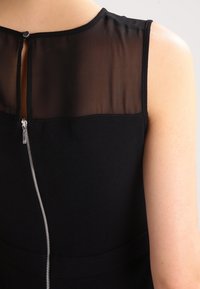 Vestido negro con un panel superior transparente, un cierre de cremallera en la parte posterior y una cintura ajustada. La tela tiene una textura suave y mate.