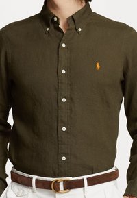 Muž v tmavě olivové košili na knoflíky s oranžovým vyšívaným logem, bílých kalhotách a hnědém semišovém pásku se zlatou přezkou.