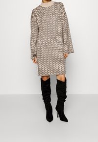 Abito lavorato a maglia beige con un motivo a onde nere. Maniche lunghe, scollo rotondo e lunghezza fino al ginocchio. Abbinato a stivali alti neri e morbidi.