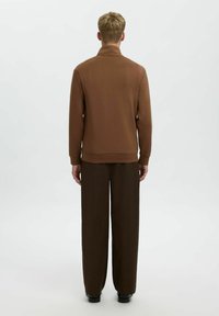 Brauner Pullover mit hohem Kragen und gerippten Bündchen, kombiniert mit passenden, lässig geschnittenen braunen Hosen, von hinten gesehen. Glatte Textur.
