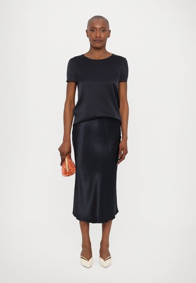 Črn satenski top s kratkimi rokavi v kombinaciji s sijočo črno midi krilo. Dodatki vključujejo inkrustirano oranžno torbico in bež čevlje s koničastim prstom.