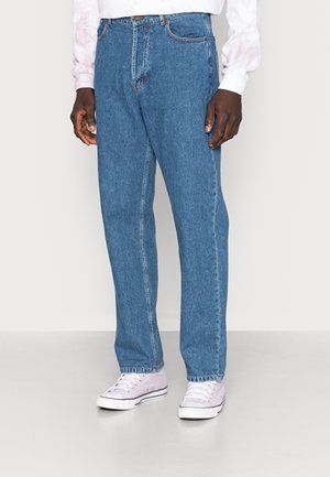 Person trägt blaue Jeans mit geradem Bein, weiße und pinke Sneaker und ein langärmliges weiß-pinkes Hemd, steht auf einem schlichten Boden.