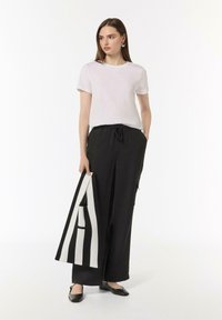 Weißes kurzärmliges T-Shirt, schwarze weite Hose mit elastischem Bund, hält eine schwarz-weiße gestreifte Tasche, kombiniert mit schwarzen Ballerinas.