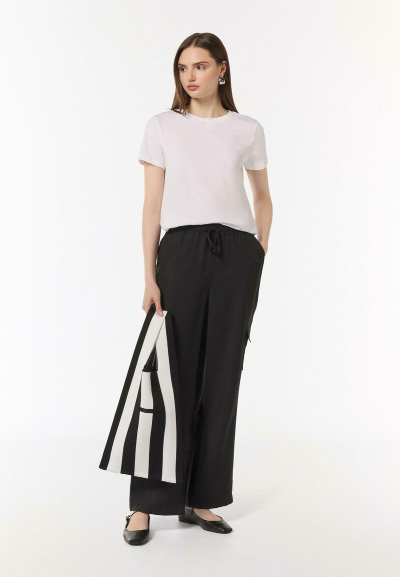 Weißes kurzärmliges T-Shirt, schwarze weite Hose mit elastischem Bund, hält eine schwarz-weiße gestreifte Tasche, kombiniert mit schwarzen Ballerinas.