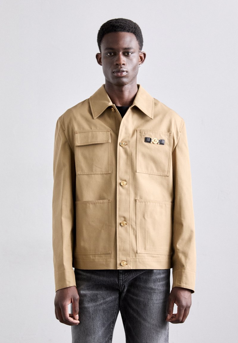 Tan cotton jacket with a collar, button closure, four front pockets, and a decorative hardware detail on the chest. Worn with gray jeans.