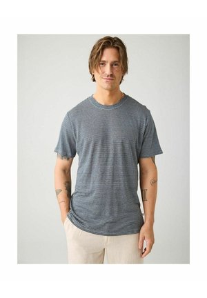 Man met halflang haar, gekleed in een zwart-wit gestreept t-shirt en lichte broek, staand met handen in de zakken.