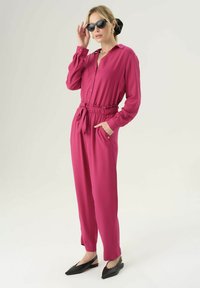 Jumpsuit lungo fucsia con maniche lunghe, chiusura a bottoni sul davanti, vita cinchata, tasche laterali e gambe tapered, abbinato a ballerine nere con punta a punta.