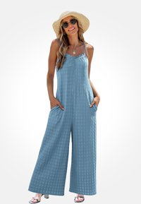 Blauwe jumpsuit met patroon en wijde pijpen, gemaakt van een textuurstof, met verstelbare banden en zijzakken. Gecombineerd met een strohoed en een zonnebril.