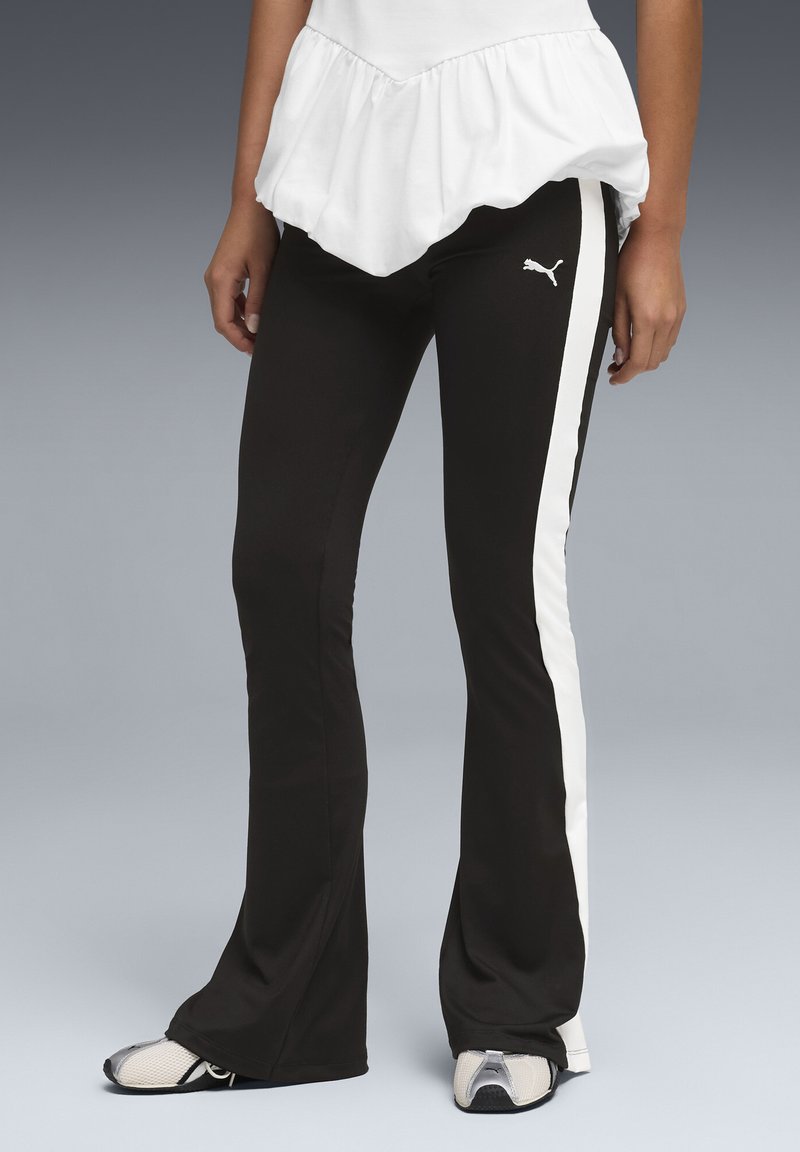 Άτομο που φοράει μαύρο καμπανέ παντελόνι με λευκές πλαϊνές ρίγες και λογότυπο Puma, συνδυασμένο με λευκά αθλητικά παπούτσια και λευκό τοπ με βολάν.