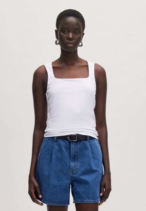 Weißes Tanktop mit quadratischem Ausschnitt, kombiniert mit high-waisted blauen Jeansshorts und einem schwarzen Gürtel. Das Model steht vor einem hellgrauen Hintergrund.