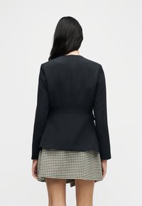 Μαύρο, κομψό σακάκι με μακριά μανίκια, εφαρμοστό και με ζώνη στη μέση που δημιουργεί πεπλό. Φορεμένο πάνω από κοντή φούστα με μοτίβο καρό κουκουνάρι.