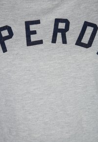 Maglietta grigia con lettere in blocco blu scuro che scrivono "SUPERDRY" sul petto. Tessuto in cotone morbido con un design a girocollo standard.