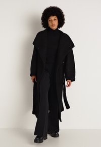 Cappotto nero texturizzato con ampi revers, bottoni e silhouette lunga, indossato sopra un dolcevita nero e pantaloni neri a zampa.