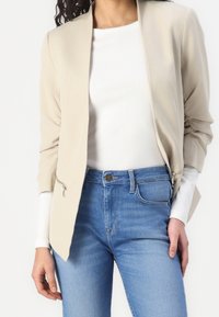 Beżowa marynarka o dopasowanym kroju, z marszczonymi detalami rękawów i zapinanymi kieszeniami, noszona na białej koszuli i niebieskich dżinsach. Gładka tekstura.