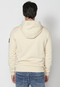 Sudadera con capucha de color crema claro, hecha de una tela suave. Presenta un bolsillo tipo canguro y puños acanalados. Se lleva con jeans azules.