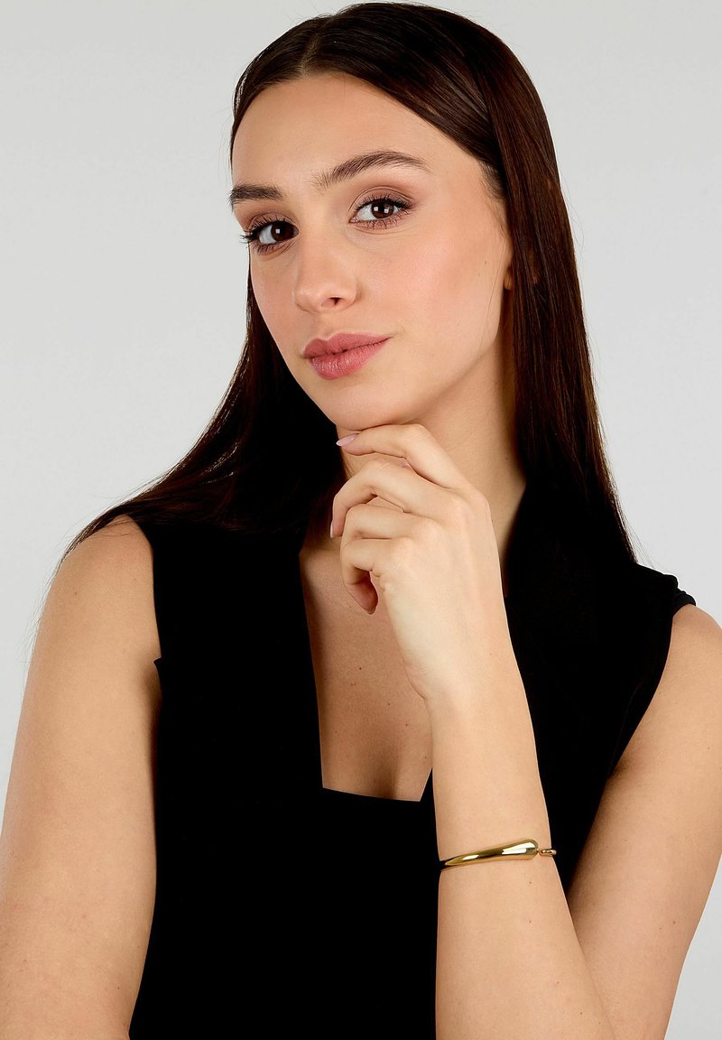 Braccialetto bangle in metallo dorato indossato su un polso, caratterizzato da un design liscio e curvato. Lo sfondo è neutro, evidenziando la lucentezza del bracciale.