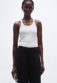 Fehér pamut trikó, testhezálló szabással, magas derekú, fekete nadrággal, amelyek redőzöttek. A modell egy kis fekete táskát tart.