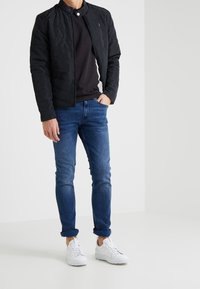 Czarna pikowana kurtka z kołnierzem stojącym, nałożona na ciemną bluzkę, w połączeniu z niebieskimi dopasowanymi dżinsami i białymi trampkami.