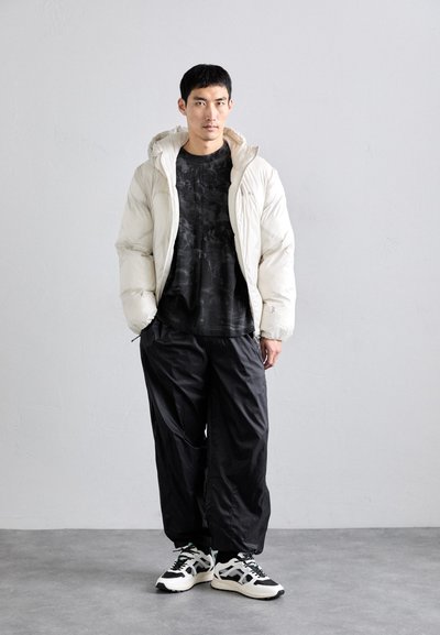 Witte pufferjack met capuchon, zwart met patroon shirt, losse zwarte broek en zwart-witte sneakers met groene accenten.