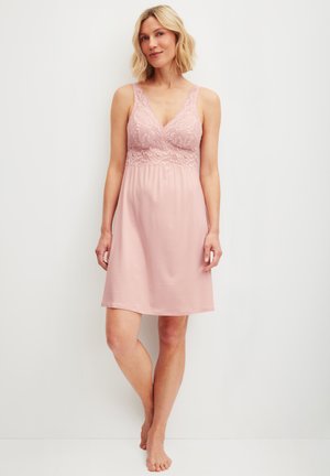 Frau steht barfuß und trägt ein ärmelloses pinkfarbenes Nachthemd mit Spitzenoberteil und V-Ausschnitt vor einem schlichten hellen Hintergrund.