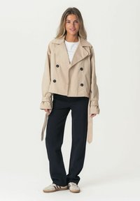 Trench coat beige cropped con bottoni neri doppi, ampie revers, e dettagli delle maniche legati, abbinato a pantaloni scuri e sneakers.
