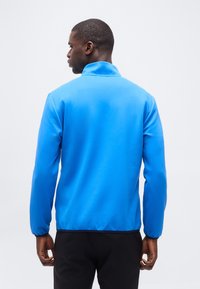 Άνδρας που φοράει φωτεινό μπλε μπουφάν με μακριά μανίκια  και ψηλό γιακά, γυρισμένος με την πλάτη προς το μέρος μας, σε απλό ανοιχτόχρωμο φόντο.