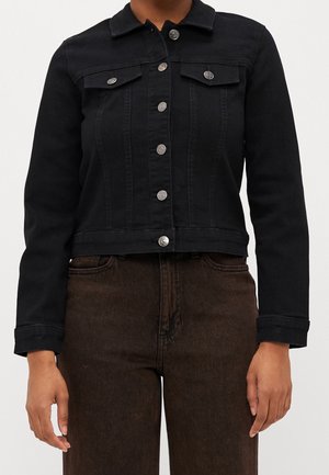 Personne portant une veste en jean noire boutonnée avec poches à rabat et un jean marron taille haute, debout devant un fond uni.