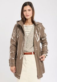 Giacca impermeabile marrone lucida con cappuccio, multiple tasche e chiusure a scatto, indossata sopra un cardigan grigio e una maglietta a righe.