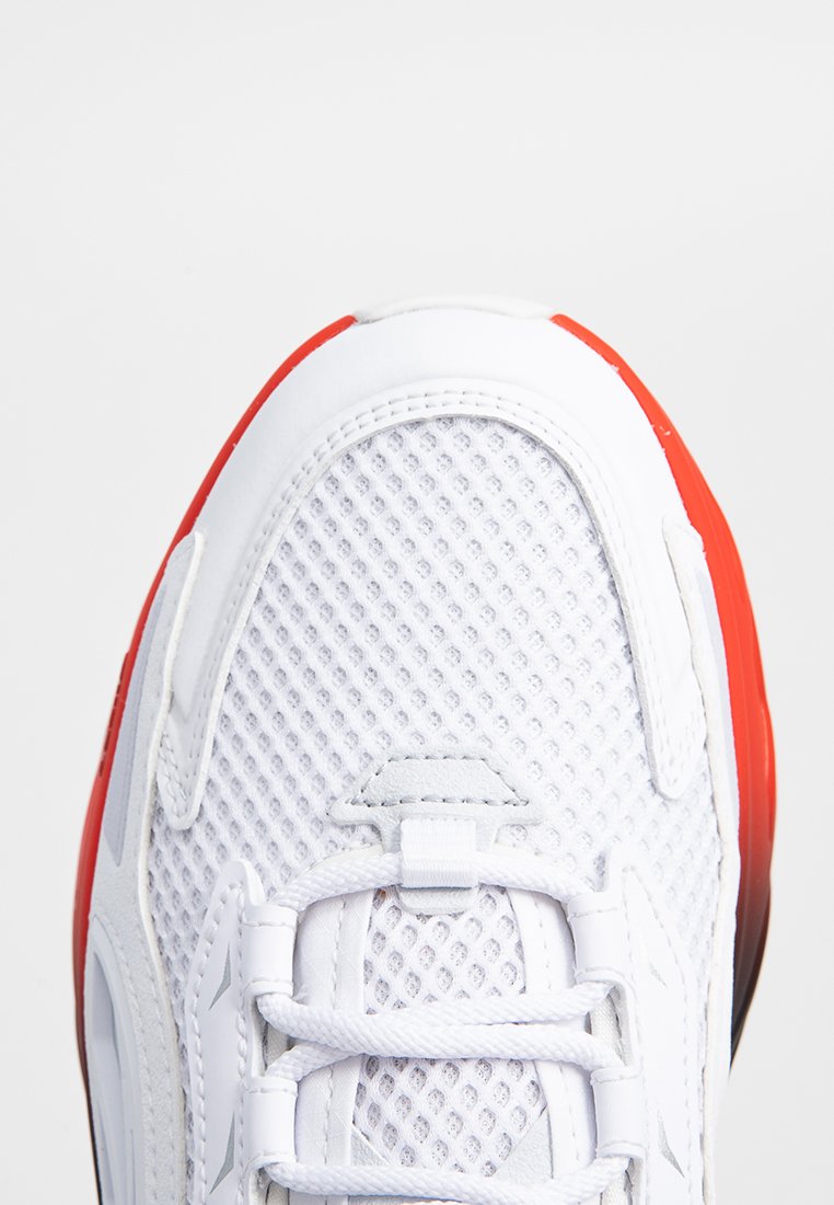 Baskets blanches avec une tige en mesh, semelle en caoutchouc rouge et lacets blancs. Les détails notables incluent un motif texturé sur la tige.