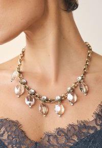 Gold chain necklace featuring faceted clear crystals and round metallic accents, with a layered design and drop elements for added detail.