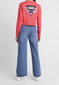 Rotes Langarmhemd mit einem schwarzen Dreieckslogo auf dem Rücken, kombiniert mit lockeren blauen Jeans und weißen Sneakers.