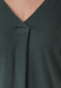 Blouse en tricot vert foncé avec un col en V et un subtil détail de pli au centre devant. Texture douce, tissu léger.