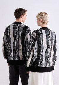 Knit sweater in black and white with a textured geometric pattern, featuring a ribbed black hem and cuffs, worn by two figures from the back.