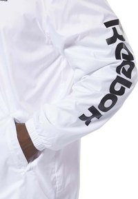 Giacca leggera bianca con polsini elasticizzati, caratterizzata da un grande logo nero "Reebok" lungo la manica. Include tasche laterali.