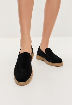 Black suede slip-on shoes with a rounded toe and a beige rubber sole. Features a smooth upper and minimal stitching details.