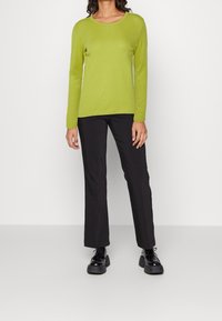 Maglione verde a maniche lunghe con scollo rotondo, abbinato a pantaloni neri a zampa d'elefante e scarpe nere spesse, in piedi davanti a uno sfondo neutro.