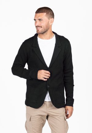 Cardigan nero con colletto, chiusura anteriore con bottoni e due tasche frontali, indossato sopra una camicia bianca. Tessuto a coste tipo fisherman, design casual.