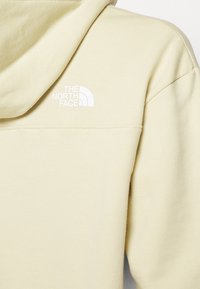 Ljusgul hoodie i mjukt tyg, med en vit logotyp av The North Face på baksidan nära halsringningen.