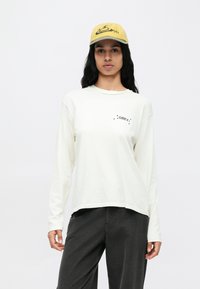 Crèmekleurig T-shirt met lange mouwen en een ronde hals, met een zwart "OBEY"-logo, gecombineerd met grijze ribfluwelen broek en een mosterdgele pet.