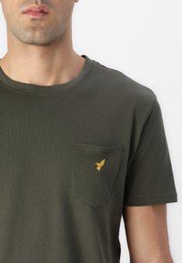 Homme portant un t-shirt vert foncé avec un petit oiseau jaune brodé sur la poche poitrine gauche, sur un fond blanc uni.